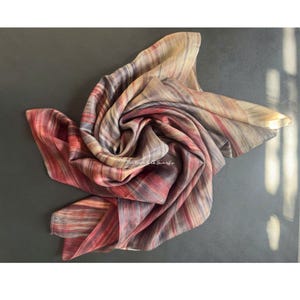 May include: A square silk scarf featuring a multi-coloured abstract pattern with vertical streaks of red, deep purple, grey, and beige tones. The fabric is gathered in the centre, creating soft folds and ripples. The text "Main Road Silk Scarfs" is visible.
