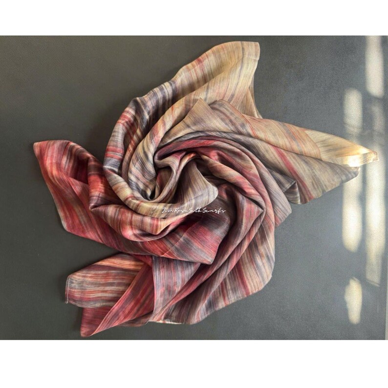 May include: A square silk scarf featuring a multi-coloured abstract pattern with vertical streaks of red, deep purple, grey, and beige tones. The fabric is gathered in the centre, creating soft folds and ripples. The text "Main Road Silk Scarfs" is visible.