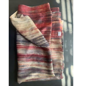 May include: A rectangular scarf featuring a horizontal striped pattern in shades of deep red, muted purple, and cream. The fabric has a subtle sheen and a visible weave texture. A small white rectangular tag is attached to the side edge of the scarf.