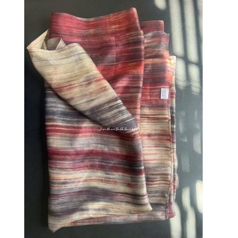 May include: A rectangular scarf featuring a horizontal striped pattern in shades of deep red, muted purple, and cream. The fabric has a subtle sheen and a visible weave texture. A small white rectangular tag is attached to the side edge of the scarf.
