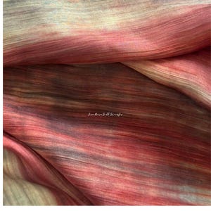 May include: A textured silk scarf featuring a gradient of blended colours including deep red, muted pink, and soft beige tones. The fabric displays horizontal striations and folds throughout, with the text "Jim Rose Silk Scarfs" printed in white.