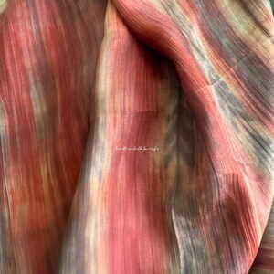 May include: A lightweight fabric scarf featuring a vertical gradient pattern in shades of coral, peach, and muted grey. The material has a visible fine-grained texture and includes the text Kim Rose Silk Scarfs printed in white in the centre.