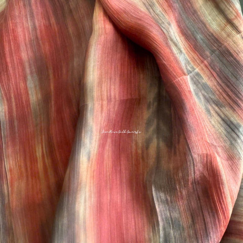May include: A lightweight fabric scarf featuring a vertical gradient pattern in shades of coral, peach, and muted grey. The material has a visible fine-grained texture and includes the text Kim Rose Silk Scarfs printed in white in the centre.