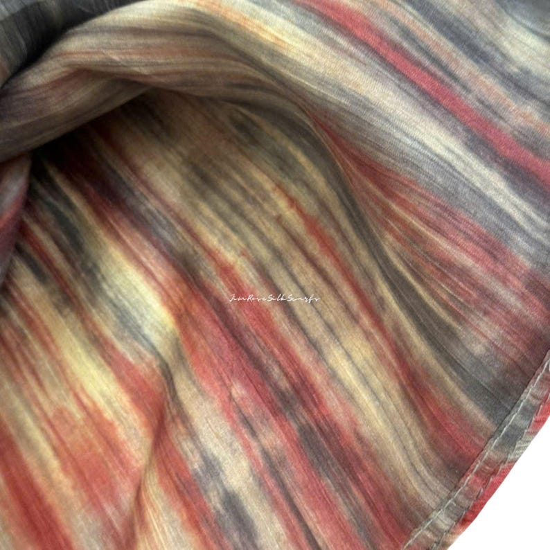 May include: A lightweight scarf featuring a vertical gradient pattern in shades of charcoal grey, muted yellow, and deep red. The fabric displays a subtle, fine-textured weave with a soft sheen. The text "June Rose Silk Scarfs" is printed in white.