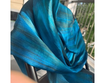 May include: A teal and blue scarf with black stripes, draped over a chair. The scarf's fabric has a soft texture and a flowing design. The colour palette includes shades of teal and blue, creating a visually appealing accessory.