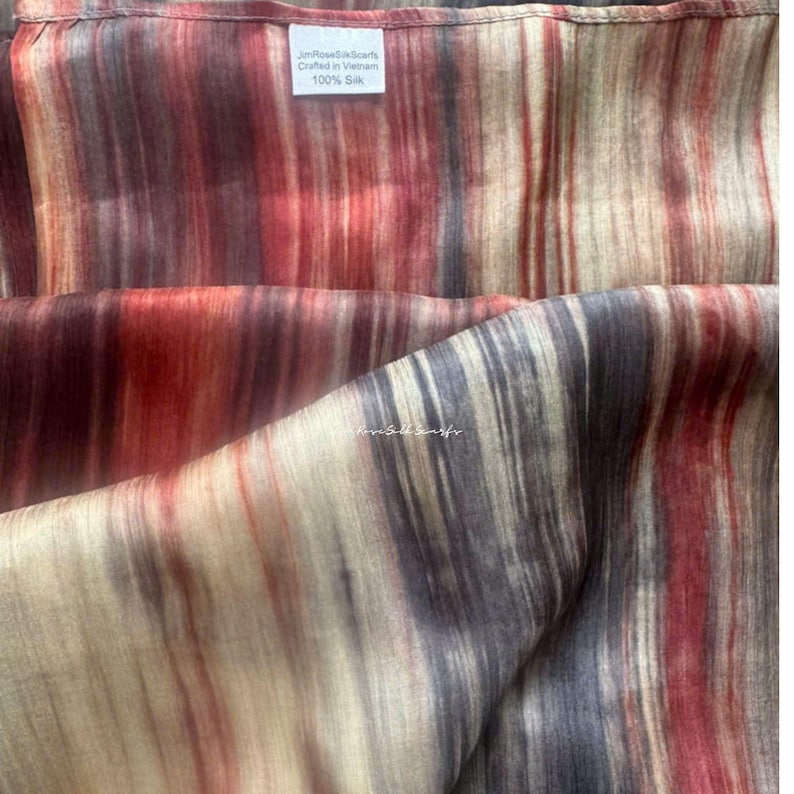 May include: A 100% silk scarf featuring a vertical striped pattern in shades of deep red, dark grey, and cream. A white rectangular fabric label is sewn onto the top edge, displaying the text JimRoseSilkScarfs, Crafted in Vietnam, 100% Silk.