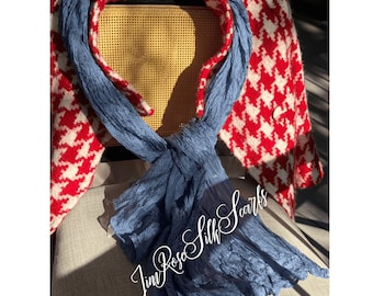 May include: A blue silk scarf is knotted in the center, with a red and white houndstooth jacket in the background. The scarf is draped over a light gray surface. The text "Jim Rose Silk Scarfs" is in the lower right corner.