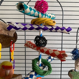 May include: A colourful bird toy hanging in a cage. The toy is made of woven straw, wood, and rope. It features a variety of colours, including blue, green, purple, red, yellow, and pink. The toy is designed to provide entertainment and enrichment for birds.