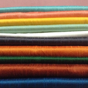 May include: A collection of fabric rolls in various colours, including teal, orange, yellow, green, white, navy, and rust. The material has a sheen, possibly silk. The rolls are stacked, displaying the range of colours available. The fabric is likely for sewing.
