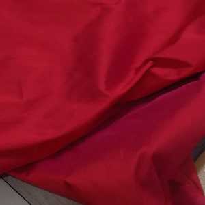 Può includere: Un primo piano di un tessuto rosso con una texture liscia. Il tessuto è piegato a metà, creando un drappeggio morbido e fluido.