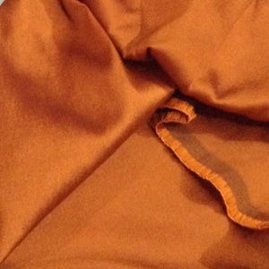 Pode incluir: Close-up de um tecido dobrado, cor de ferrugem. O tecido tem uma textura macia e é drapeado de forma a criar sombras e realces. A borda do tecido é visível, com uma borda texturizada contrastante.