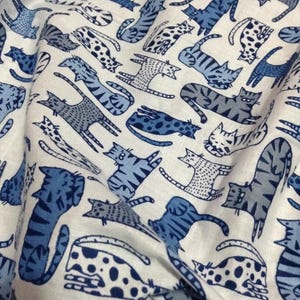 Peut inclure: Un tissu blanc orné d'un motif répétitif de chats dans diverses nuances de bleu et de gris. Les chats sont représentés dans différentes postures, notamment assis, étirés et allongés, avec des motifs variés comme des rayures et des taches.