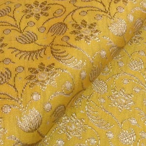Peut inclure: Gros plan sur un tissu jaune avec un motif floral doré complexe. Le motif présente des fleurs stylisées, des feuilles et des lignes tourbillonnantes, créant un aspect riche et texturé. Le tissu est plié, révélant le tissage et le lustre détaillés.