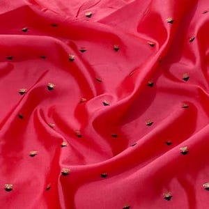 May include: A vibrant red fabric featuring a repeating pattern of small, embroidered gold-colored floral motifs scattered across the surface. The material is draped in soft folds, highlighting the smooth texture and the metallic sheen of the thread.