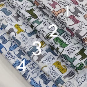 Op de afbeelding: Stalen stof met kattenpatronen in diverse kleuren. De ontwerpen omvatten katten met strepen, stippen en effen kleuren, tegen een witte achtergrond. De stof is gevouwen en toont verschillende patronen.