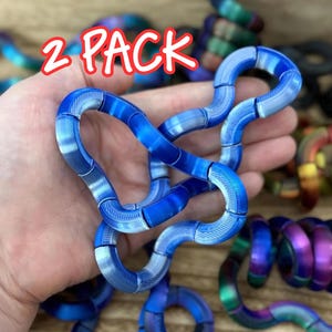 Puede incluir: Un paquete de 2 juguetes antiestr&eacute;s azules formados por segmentos curvos interconectados con una superficie texturizada. Los juguetes son flexibles y pueden manipularse en varias formas. Se ven otros juguetes multicolores y negros.