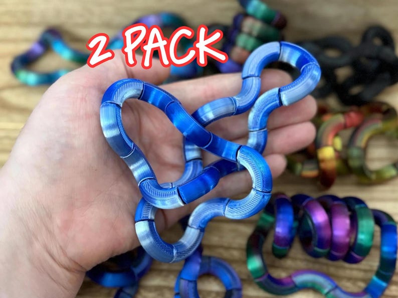 Puede incluir: Un paquete de 2 juguetes antiestr&eacute;s azules formados por segmentos curvos interconectados con una superficie texturizada. Los juguetes son flexibles y pueden manipularse en varias formas. Se ven otros juguetes multicolores y negros.