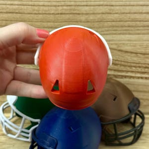 Puede incluir: Un peque&ntilde;o casco de f&uacute;tbol americano de pl&aacute;stico naranja con una m&aacute;scara facial blanca y dos orificios de ventilaci&oacute;n triangulares en la parte superior. El casco se sostiene frente a otros cascos en miniatura verdes, azules y marrones.