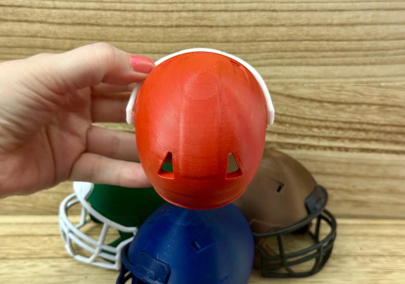 Puede incluir: Un peque&ntilde;o casco de f&uacute;tbol americano de pl&aacute;stico naranja con una m&aacute;scara facial blanca y dos orificios de ventilaci&oacute;n triangulares en la parte superior. El casco se sostiene frente a otros cascos en miniatura verdes, azules y marrones.