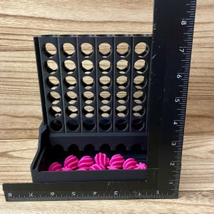 May include: A black plastic vertical grid game board with seven columns and six rows of circular openings. A collection of small, bright pink spherical game pieces rests in the base tray. A black L-shaped ruler shows a height of 20 cm.