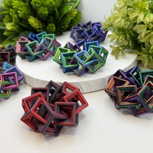 Pode incluir: Uma cole&ccedil;&atilde;o de esculturas geom&eacute;tricas coloridas. Cada escultura &eacute; composta por molduras quadradas interconectadas, exibindo um espectro de cores, incluindo vermelho, verde, azul e roxo. As esculturas est&atilde;o dispostas sobre uma superf&iacute;cie branca com folhagem verde ao fundo.