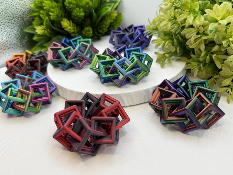 Pode incluir: Uma cole&ccedil;&atilde;o de esculturas geom&eacute;tricas coloridas. Cada escultura &eacute; composta por molduras quadradas interconectadas, exibindo um espectro de cores, incluindo vermelho, verde, azul e roxo. As esculturas est&atilde;o dispostas sobre uma superf&iacute;cie branca com folhagem verde ao fundo.