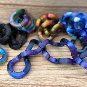 Puede incluir: Una colecci&oacute;n de juguetes fidget articulados hechos de segmentos curvos interconectados. Las piezas presentan acabados met&aacute;licos iridiscentes en tonos azul, morado, verde y naranja, junto con varios segmentos negros mate texturizados.