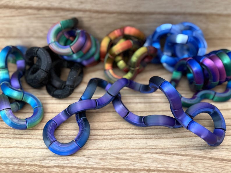Puede incluir: Una colecci&oacute;n de juguetes fidget articulados hechos de segmentos curvos interconectados. Las piezas presentan acabados met&aacute;licos iridiscentes en tonos azul, morado, verde y naranja, junto con varios segmentos negros mate texturizados.