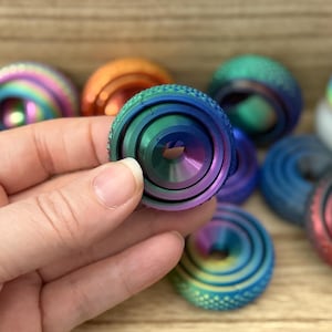May include: A hand holds a multi-coloured, iridescent metal fidget spinner. Other spinners in various colours and designs are in the background. The fidget spinner has a circular design with concentric rings and a central hole.