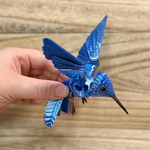 May include: A detailed blue hummingbird sculpture with outstretched wings. The bird has intricate feather detailing and a long, slender beak. The sculpture is held against a wooden background, showcasing its vibrant colour and design.