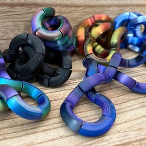 Puede incluir: Una colecci&oacute;n de juguetes sensoriales articulados formados por segmentos curvos interconectados. Las piezas presentan varios acabados, incluyendo negro mate, arco&iacute;ris iridiscente, azul met&aacute;lico y tonos degradados naranja-amarillo.