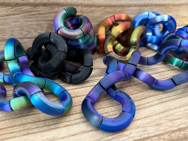 Puede incluir: Una colecci&oacute;n de juguetes sensoriales articulados formados por segmentos curvos interconectados. Las piezas presentan varios acabados, incluyendo negro mate, arco&iacute;ris iridiscente, azul met&aacute;lico y tonos degradados naranja-amarillo.