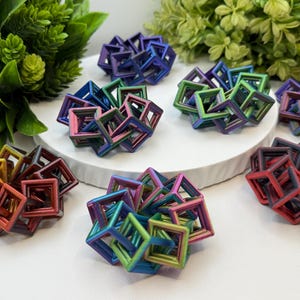 Pode incluir: Uma cole&ccedil;&atilde;o de esculturas geom&eacute;tricas coloridas em v&aacute;rios tons de vermelho, laranja, verde, azul e roxo. As esculturas est&atilde;o dispostas sobre uma superf&iacute;cie branca com folhagem verde ao fundo. Os objetos s&atilde;o feitos de molduras quadradas interconectadas.