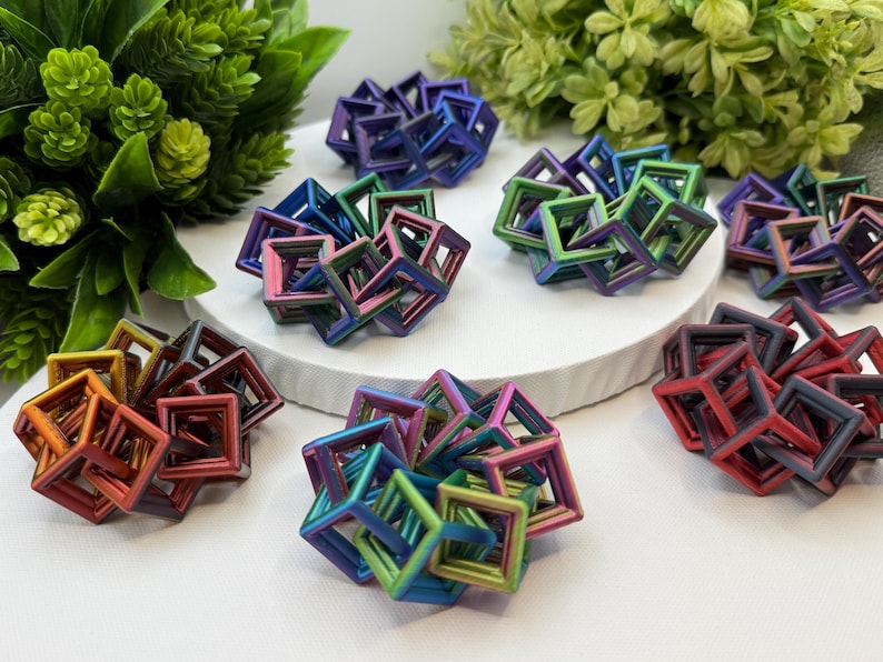 Pode incluir: Uma cole&ccedil;&atilde;o de esculturas geom&eacute;tricas coloridas em v&aacute;rios tons de vermelho, laranja, verde, azul e roxo. As esculturas est&atilde;o dispostas sobre uma superf&iacute;cie branca com folhagem verde ao fundo. Os objetos s&atilde;o feitos de molduras quadradas interconectadas.