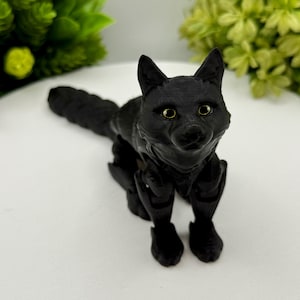 May include: A matte black articulated fox figurine with a textured surface, pointed ears, and detailed facial features. The fox has light-colored eyes and is posed in a standing position with its long, segmented tail extending behind its body.