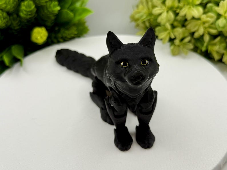 May include: A matte black articulated fox figurine with a textured surface, pointed ears, and detailed facial features. The fox has light-colored eyes and is posed in a standing position with its long, segmented tail extending behind its body.