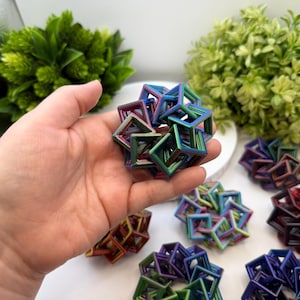 Pode incluir: Uma m&atilde;o segurando uma escultura c&uacute;bica geom&eacute;trica multicolorida. V&aacute;rias esculturas semelhantes em v&aacute;rias cores, incluindo vermelho, azul e verde, est&atilde;o dispostas em uma superf&iacute;cie branca. Plantas verdes e frondosas ao fundo.