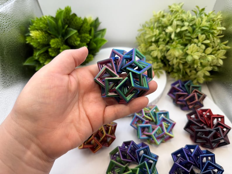 Pode incluir: Uma m&atilde;o segurando uma escultura c&uacute;bica geom&eacute;trica multicolorida. V&aacute;rias esculturas semelhantes em v&aacute;rias cores, incluindo vermelho, azul e verde, est&atilde;o dispostas em uma superf&iacute;cie branca. Plantas verdes e frondosas ao fundo.