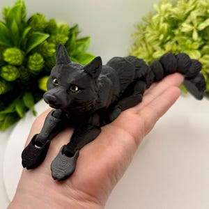 Puede incluir: Una figura de lobo negro articulada con ojos dorados. El lobo está tumbado, con las patas y la cola en posición relajada. La figura es detallada y parece estar impresa en 3D. El fondo presenta follaje verde.