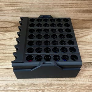 May include: A black plastic storage organiser featuring a grid of circular holes arranged in rows and columns. The unit has a solid base and a side panel with a series of semi-circular cutouts along the edge, designed for holding small items.