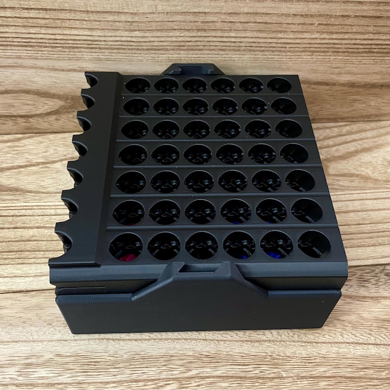 May include: A black plastic storage organiser featuring a grid of circular holes arranged in rows and columns. The unit has a solid base and a side panel with a series of semi-circular cutouts along the edge, designed for holding small items.