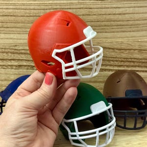 Puede incluir: Una colecci&oacute;n de cascos de f&uacute;tbol americano en miniatura de varios colores, incluyendo rojo, azul, verde y marr&oacute;n. Cada casco cuenta con una m&aacute;scara protectora, siendo la m&aacute;scara del casco rojo y del casco verde de color blanco.