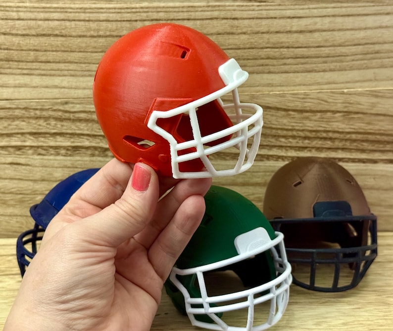 Puede incluir: Una colecci&oacute;n de cascos de f&uacute;tbol americano en miniatura de varios colores, incluyendo rojo, azul, verde y marr&oacute;n. Cada casco cuenta con una m&aacute;scara protectora, siendo la m&aacute;scara del casco rojo y del casco verde de color blanco.