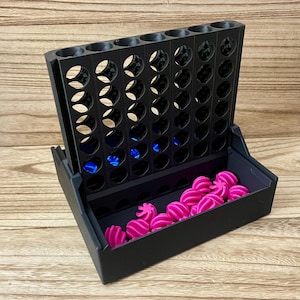 May include: A black plastic vertical grid game board with seven columns and six rows. The base features a storage tray containing multiple small, bright pink, spiraled, star-shaped game pieces, with a few blue pieces visible inside the grid slots.