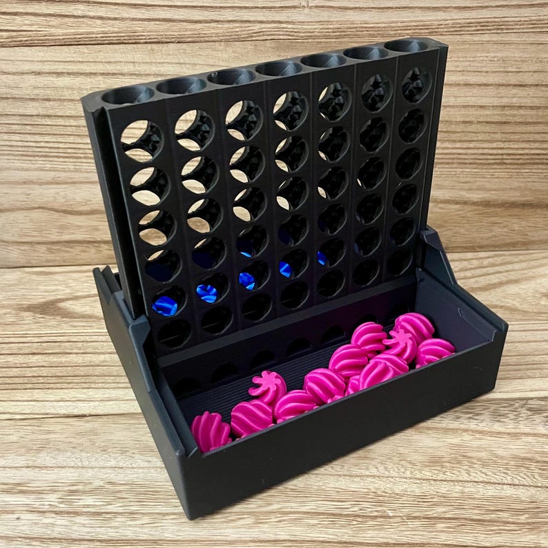 May include: A black plastic vertical grid game board with seven columns and six rows. The base features a storage tray containing multiple small, bright pink, spiraled, star-shaped game pieces, with a few blue pieces visible inside the grid slots.