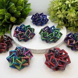 Pode incluir: Uma cole&ccedil;&atilde;o de esculturas geom&eacute;tricas coloridas em v&aacute;rios tons de vermelho, azul, verde e roxo. Cada escultura &eacute; composta por formas c&uacute;bicas interconectadas. As esculturas est&atilde;o dispostas sobre uma superf&iacute;cie branca, com folhagem verde ao fundo.