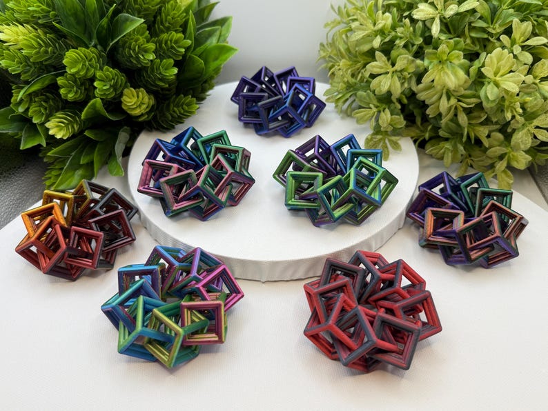 Pode incluir: Uma cole&ccedil;&atilde;o de esculturas geom&eacute;tricas coloridas em v&aacute;rios tons de vermelho, azul, verde e roxo. Cada escultura &eacute; composta por formas c&uacute;bicas interconectadas. As esculturas est&atilde;o dispostas sobre uma superf&iacute;cie branca, com folhagem verde ao fundo.
