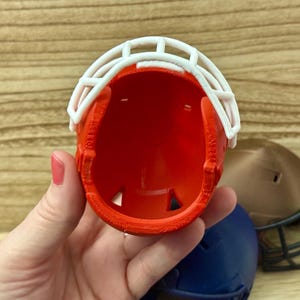 Puede incluir: Un casco de f&uacute;tbol americano en miniatura de color naranja con una m&aacute;scara facial blanca. El casco es hueco y muestra una superficie interior lisa. Otros cascos en miniatura en azul oscuro y marr&oacute;n son parcialmente visibles al fondo.