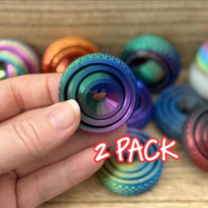 May include: A collection of circular, multi-colored metal fidget spinners with a textured, knurled outer edge and a smooth, concave center. The items feature a vibrant gradient of blue, green, and purple hues. The text 2 PACK is visible.