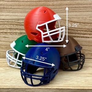Puede incluir: Una colecci&oacute;n de cuatro cascos de f&uacute;tbol americano en miniatura de colores rojo, verde, azul y marr&oacute;n, cada uno con una rejilla protectora. Los cascos miden 8,26 cm de alto, 10,16 cm de ancho y 8,26 cm de profundidad, seg&uacute;n las flechas.